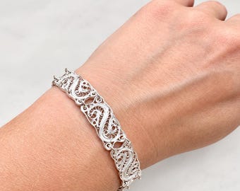 May include: A silver bracelet featuring a series of interconnected rectangular links, each displaying an intricate filigree pattern of swirling wirework. The metallic links are joined together by small circular rings, creating a flexible band.