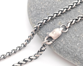 May include: A silver-toned wheat chain necklace featuring a polished lobster claw clasp. The metal links have a darkened, oxidised finish within the crevices, creating a textured appearance along the length of the chain.