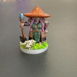May include: A painted miniature figurine of a character wearing a large orange mushroom cap as a hat, featuring pink braided hair, a green tunic, and brown armor. The figure holds a wooden staff with a blue crystal and stands on a base.