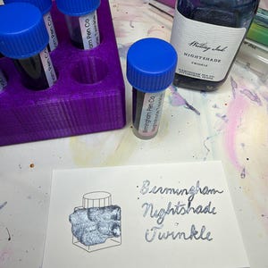 May include: A collection of Birmingham Pen Co. Nightshade Twinkle writing ink in a large glass bottle and three small sample vials with blue caps, stored in a purple plastic holder, alongside a white card with the handwritten text Birmingham Nightshade Twinkle.