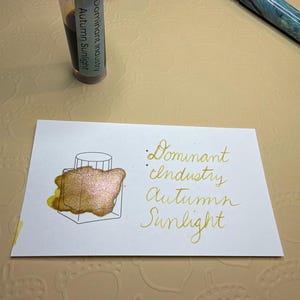 May include: A small plastic vial of Dominant Industry Autumn Sunlight ink with a purple cap, a fountain pen with a blue and green marbled barrel, and a white rectangular card featuring a gold ink swatch and the text Dominant Industry Autumn Sunlight.