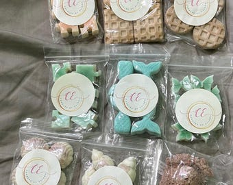 Könnte beinhalten: Neun durchsichtige Plastikbeutel mit verschiedenen Wachsschmelzstücken in Formen wie Puzzleteilen, Waffeln, Meerjungfrauenschwänzen, Schleifen und Muscheln. Jeder Beutel hat ein rundes weißes Etikett mit dem Text CE Candles by Eliana in Gold und Pink.