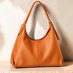 May include: A solid tan hobo-style shoulder bag with a smooth texture, a single arched handle, and a vertical seam running down the center of the front panel. The bag features a silver-toned metal buckle detail on the side strap attachment.