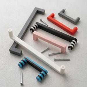 Può includere: Una collezione di maniglie per armadietti in vari colori e design, tra cui barre solide grigie, arancioni, bianche e rosa, insieme a maniglie nere con estremità a righe bianche o blu. Diverse viti di montaggio sono sparse tra gli accessori.