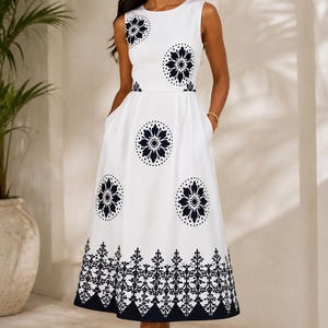 May include: A sleeveless white midi dress featuring a round neckline and a fitted bodice. The dress is decorated with dark navy blue circular floral patterns and a wide, intricate geometric border along the hemline, creating a distinct contrast.