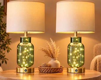 May include: A pair of table lamps featuring cylindrical green glass bases filled with warm white fairy lights, topped with off-white fabric drum shades and supported by circular gold-toned metal bases with a power button.