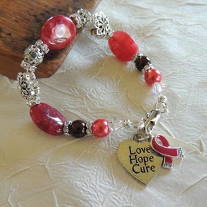 May include: A beaded bracelet featuring red and silver beads, with a heart-shaped charm that reads "Love Hope Cure" and a red ribbon. The bracelet is on a white surface.