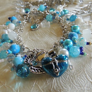 May include: A silver chain bracelet adorned with various blue and white beads, including round, faceted, and pearl-like beads. A heart-shaped charm with the word "Strength" and a blue heart-shaped locket add detail. The bracelet has a delicate, feminine aesthetic.