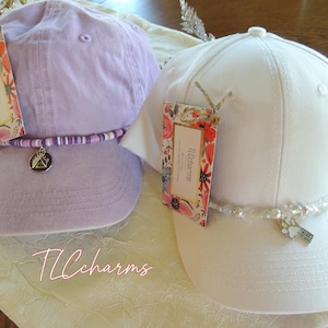 May include: Two baseball caps, one lavender and one white, each adorned with a decorative beaded band and a charm. The lavender cap has a silver charm with a mountain design, while the white cap features a paw print charm. Both caps have tags.