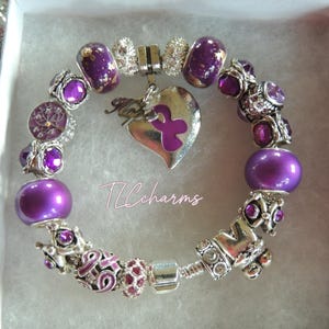May include: A silver-tone charm bracelet featuring purple beads, clear rhinestones, and a heart-shaped pendant with a purple ribbon and the word Hope. The bracelet includes various metallic spacers and beads, with the text TLCcharms visible in the center.