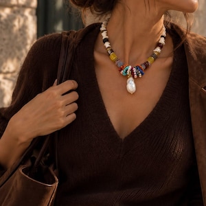May include: A beaded necklace featuring a central white baroque pearl pendant, surrounded by a mix of small, colorful beads in shades of red, blue, yellow, and white, worn with a dark brown V-neck sweater and light-colored trousers.