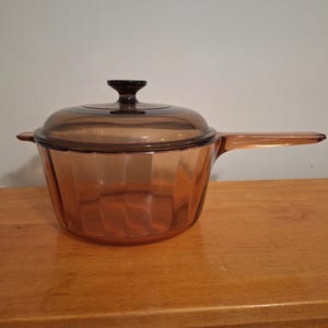 Pode incluir: Uma panela de vidro translúcido na cor âmbar com tampa a condizer. O recipiente apresenta um design canelado com estrias verticais nas laterais, uma pega longa única que se estende de um lado e um pequeno puxador central na tampa.