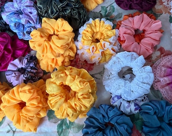 May include: A collection of fabric hair scrunchies in various colors and textures, including solid yellow, pink, dark green, and blue, alongside versions featuring white lace trim, floral patterns, and dark decorative prints.