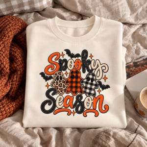 May include: A cream-colored crewneck sweatshirt featuring a graphic print with the text Spooky Season in orange and black. The design includes three ghosts with leopard print, red and black plaid, and black and white buffalo check patterns.