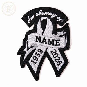 Puede incluir: Un parche bordado en blanco y negro con un diseño de cinta conmemorativa. El texto "In Memory of" está arqueado en la parte superior, con "NAME" centrado en la cinta y los años "1959" y "2026" bordados en los extremos inferiores.