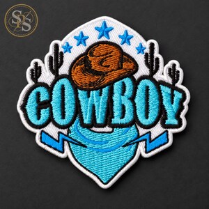 May include: An embroidered patch featuring the word COWBOY in bright blue letters with black outlines. Above the text is a brown cowboy hat, and below is a blue bandana. The design includes five blue stars and four black cactus silhouettes.