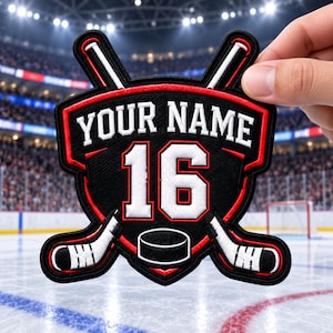 Peut inclure: Un écusson brodé noir et rouge affichant le texte YOUR NAME au-dessus du numéro 16 en blanc. Le motif comprend deux crosses de hockey croisées et un palet en bas, le tout souligné par un fil rouge et noir sur une base en forme de bouclier.