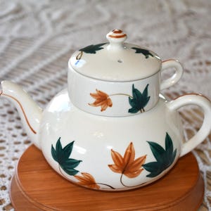 May include: White ceramic teapot with a lid and a brown wooden base. The teapot is decorated with green and brown leaves.