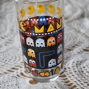 May include: A clear glass drinking glass with a Pac-Man design. The glass features a black background with yellow Pac-Man characters, blue and red maze lines, and colorful ghosts. The text "Pac-Man" is printed in yellow letters on the glass.