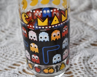 May include: A clear glass drinking glass with a Pac-Man design. The glass features a black background with yellow Pac-Man characters, blue and red maze lines, and colorful ghosts. The text "Pac-Man" is printed in yellow letters on the glass.