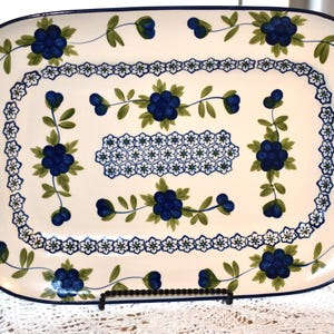 May include: An oval serving platter with a white background and a blue floral design. The platter features a dark blue border and handles. The floral pattern includes blue flowers and green leaves, with a decorative border of small blue flowers.