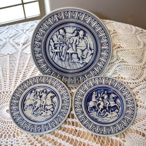 May include: Three blue and white ceramic plates with decorative borders. The plates feature scenes of knights on horseback and two men sitting at a table.