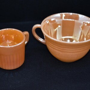 May include: Two pieces of vintage orange glassware. A small mug with a handle and vertical ridges sits next to a larger teacup with a handle and horizontal ridges. Both pieces have a glossy finish and are set against a black background.