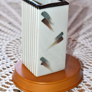 May include: A rectangular white ceramic vase with a dark brown rim and abstract brushstroke designs in green and brown. The vase is on a round, wooden base.