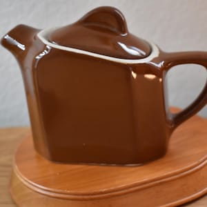 May include: A brown ceramic teapot with a lid and handle, set on a wooden stand. The teapot has a square shape with rounded edges and a white trim around the lid. The handle is a curved loop.