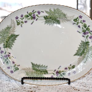 May include: Oval serving platter with a white background, adorned with a floral and fern design in green and purple. The rim has a gold trim. The platter is displayed on a black metal stand.