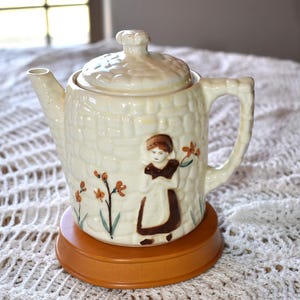 May include: A white ceramic teapot with a brick pattern and a girl in a brown dress holding flowers. The teapot has a lid and a brown wooden base.