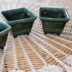 May include: Four square green ceramic planters with a decorative design. The planters have a glazed finish and are sitting on a white crocheted surface.