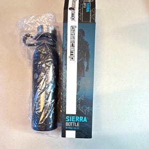 Könnte beinhalten: Eine schwarze Basecamp Sierra Flasche in einer durchsichtigen Plastiktüte neben ihrer hohen rechteckigen Verpackung. Die Schachtel zeigt das Basecamp-Logo, die Silhouette einer Person und den Text Sierra Bottle BC7056 - 24 oz. Die Flasche hat einen silbernen Verschluss.