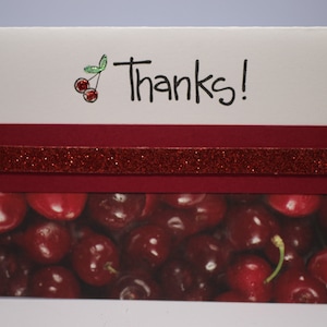 May include: A white card with the word "Thanks!" written in black ink. A small red cherry with a green stem is drawn in the top left corner. A red glitter ribbon is on top of a red background. The bottom of the card features a close-up image of a pile of red cherries.