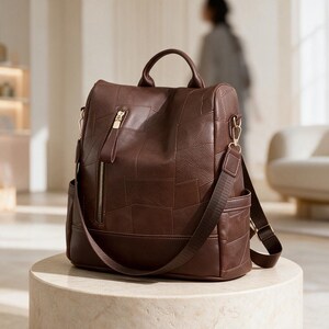 May include: A dark brown backpack featuring a patchwork design with visible stitching. The bag includes a vertical zippered front pocket with a gold-toned pull, a top handle, side pockets, and adjustable brown fabric shoulder straps with gold hardware.
