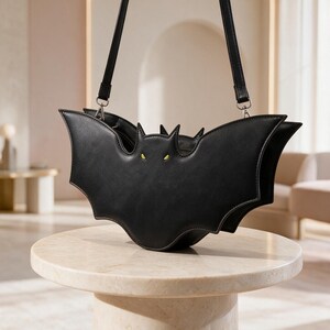 Può includere: Una borsa a tracolla nera a forma di pipistrello con finitura liscia, orecchie a punta e due piccoli occhi verde lime brillante. La borsa include una sottile tracolla nera fissata con clip in metallo color argento sulla parte superiore delle ali.