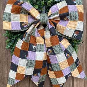 May include: A large decorative bow made of quilted fabric featuring a patchwork pattern of squares in shades of burnt orange, lavender, sage green, and cream. The fabric includes prints of small flowers, gingham checks, and leafy vines.
