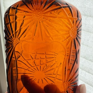 May include: An amber-colored glass bottle featuring a raised, pressed floral pattern across its surface. The bottle has a smooth, rectangular central panel and a dark, threaded cap at the top, with a translucent quality that allows light to pass through.