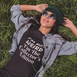 May include: A black t-shirt featuring the white text "I'M NOT WEIRD I'm The Collector's Edition" worn with a light blue denim jacket, a dark green baseball cap, and round sunglasses with dark lenses, showcasing a casual layered outfit.