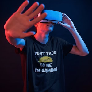 May include: A person wearing a black t-shirt with the text "Don't taco to me I'm gaming" and a cartoon taco graphic. The person is wearing a virtual reality headset and has their hand raised in front of their face.