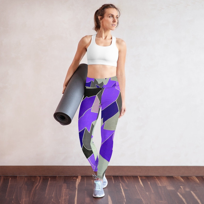 May include: A woman wearing a white sports bra and purple, black, and gray patterned yoga pants. She is holding a black yoga mat.