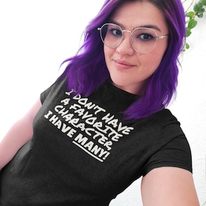 Puede incluir: Una mujer con el pelo morado y gafas lleva una camiseta negra con el texto "No tengo un personaje favorito, ¡tengo muchos!" impreso en ella.