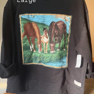 May include: A black long-sleeve sweatshirt in size Large featuring a rectangular graphic patch on the front. The patch depicts three horses grazing in a green field near a blue body of water. The sweatshirt has a crew neckline and a small white tag.