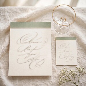 May include: A large rectangular card and a smaller rectangular card, both featuring cream-colored paper with a sage green top border and elegant gray cursive calligraphy, accompanied by a clear glass coupe with a thin gold rim.