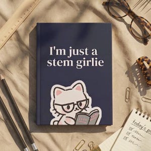 May include: A dark blue hardcover journal featuring the white text "I'm just a stem girlie" above a graphic of a white cat wearing black glasses and reading a book. The journal is positioned on a tan fabric surface with various stationery items.