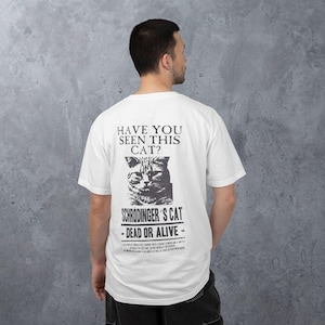 May include: A white short-sleeved t-shirt featuring a black graphic on the back. The print displays a cat illustration with the text "HAVE YOU SEEN THIS CAT?", "SCHRODINGER'S CAT", and "DEAD OR ALIVE" along with additional small printed text.