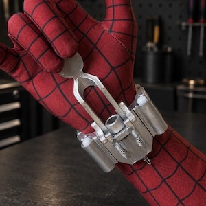May include: A silver-coloured mechanical wrist accessory featuring a central trigger mechanism and cylindrical side components, worn over a red fabric glove with a black grid pattern. The device has a metallic finish and intricate structural details.