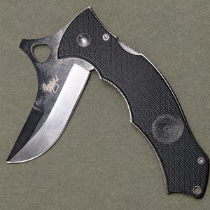 Könnte beinhalten: Ein Klappmesser mit einem schwarzen, strukturierten Griff und einer teilweise geöffneten, geschwungenen silbernen Klinge. Die Klinge weist an der Basis ein dunkles, verwittertes Finish und eine kleine eingravierte Signatur auf.