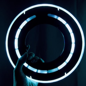 May include: A circular, black ring-shaped prop featuring a bright, glowing light blue LED strip along its outer edge. The design includes segmented light panels arranged in a ring formation, creating a futuristic appearance with high contrast.