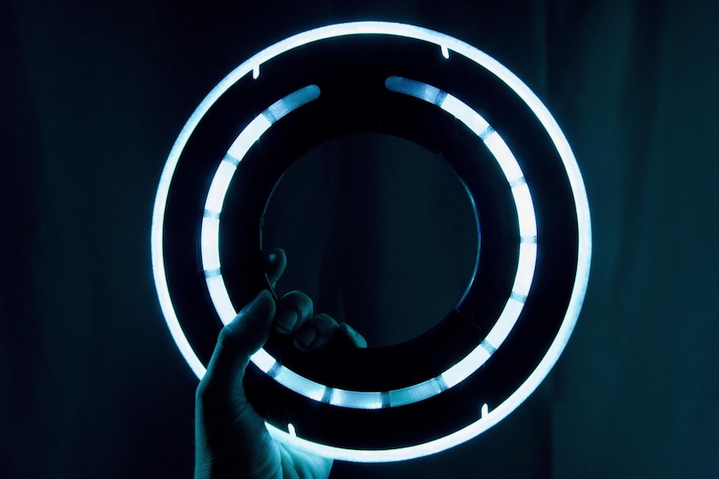 May include: A circular, black ring-shaped prop featuring a bright, glowing light blue LED strip along its outer edge. The design includes segmented light panels arranged in a ring formation, creating a futuristic appearance with high contrast.